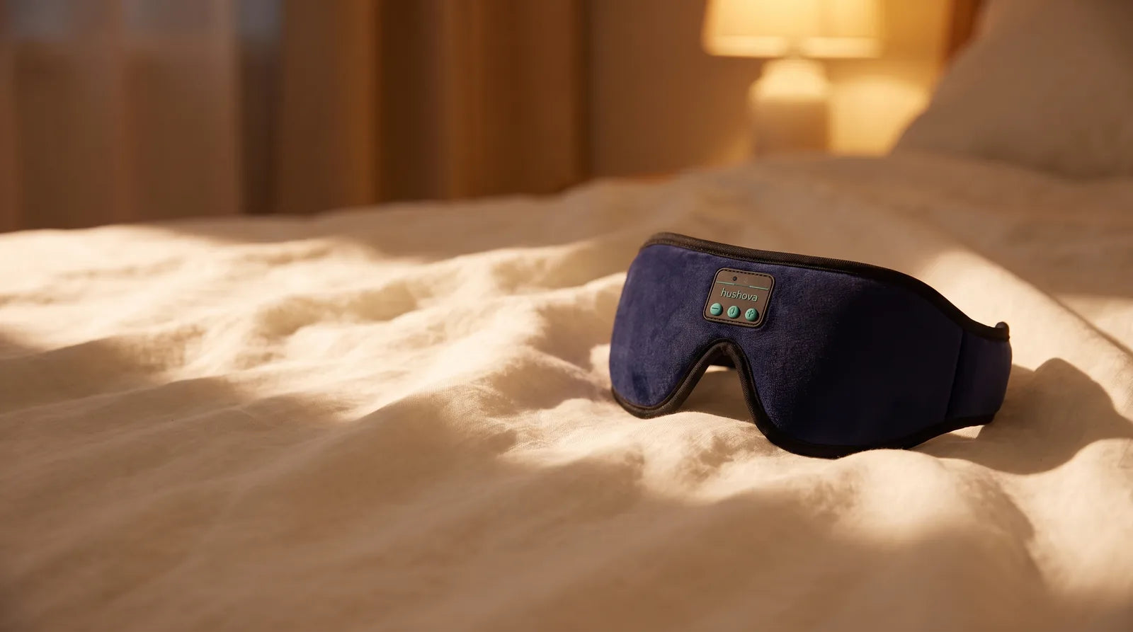 Hushova sleep mask on bedding by a bedside lamp at dusk